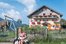 Mädchen sitzt im Garten mit einer Ziege. Im Hintergrund ein Bauernhof mit Spielplatz und den Eltern im Liegestuhl